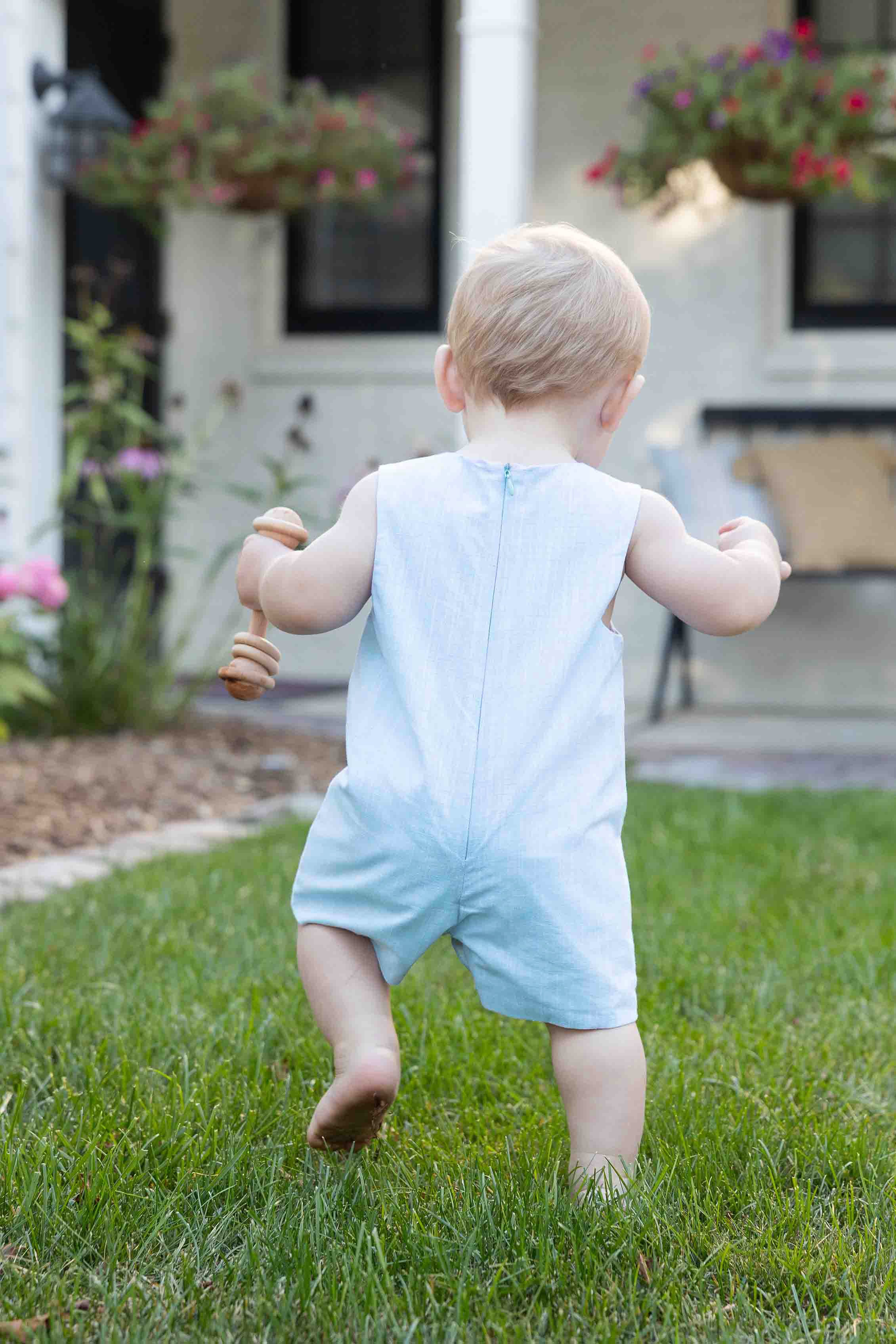Child in a light blue romper walking on grass with a house and garden in the background