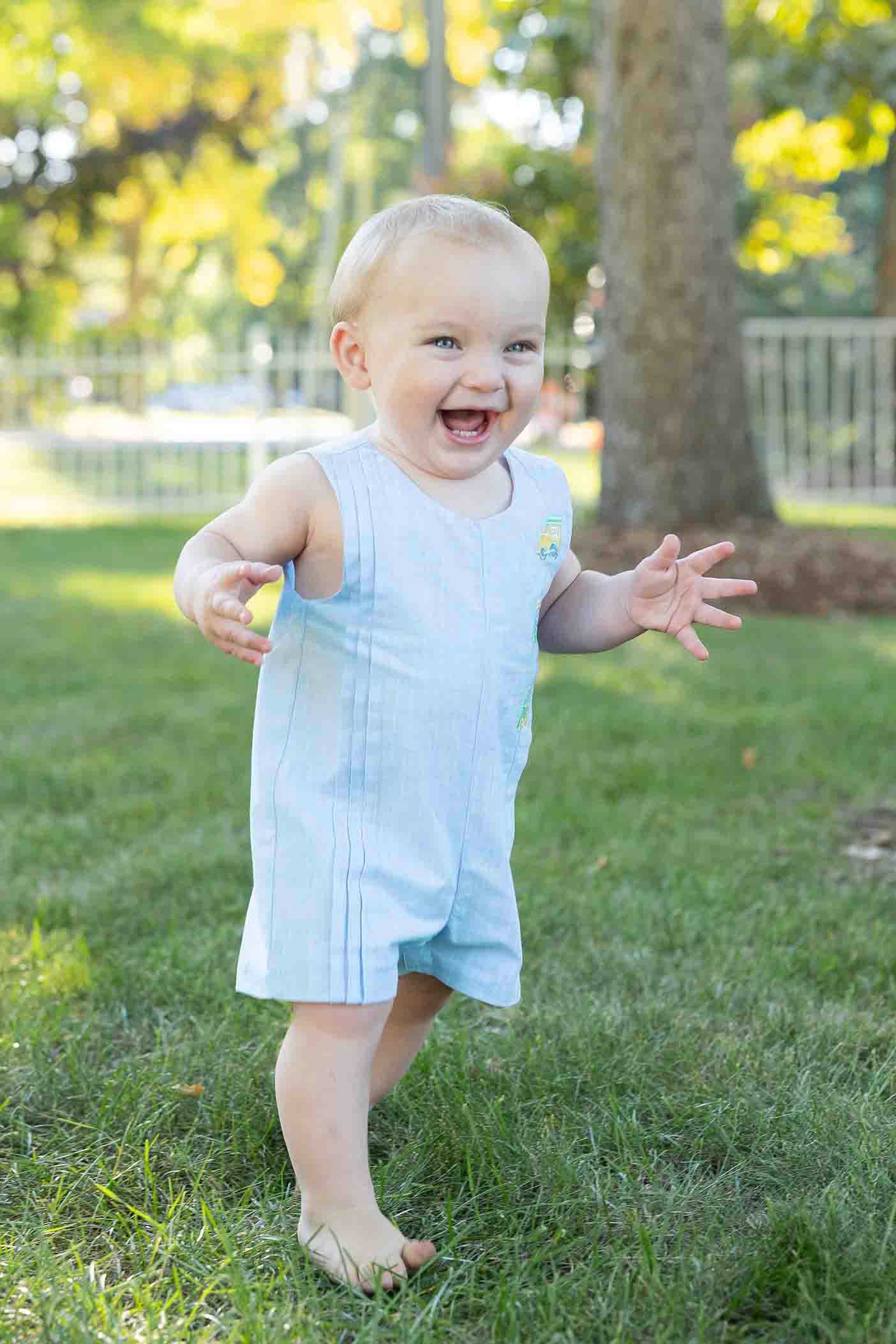 Baby in a light blue romper standing on grass with trees in the background