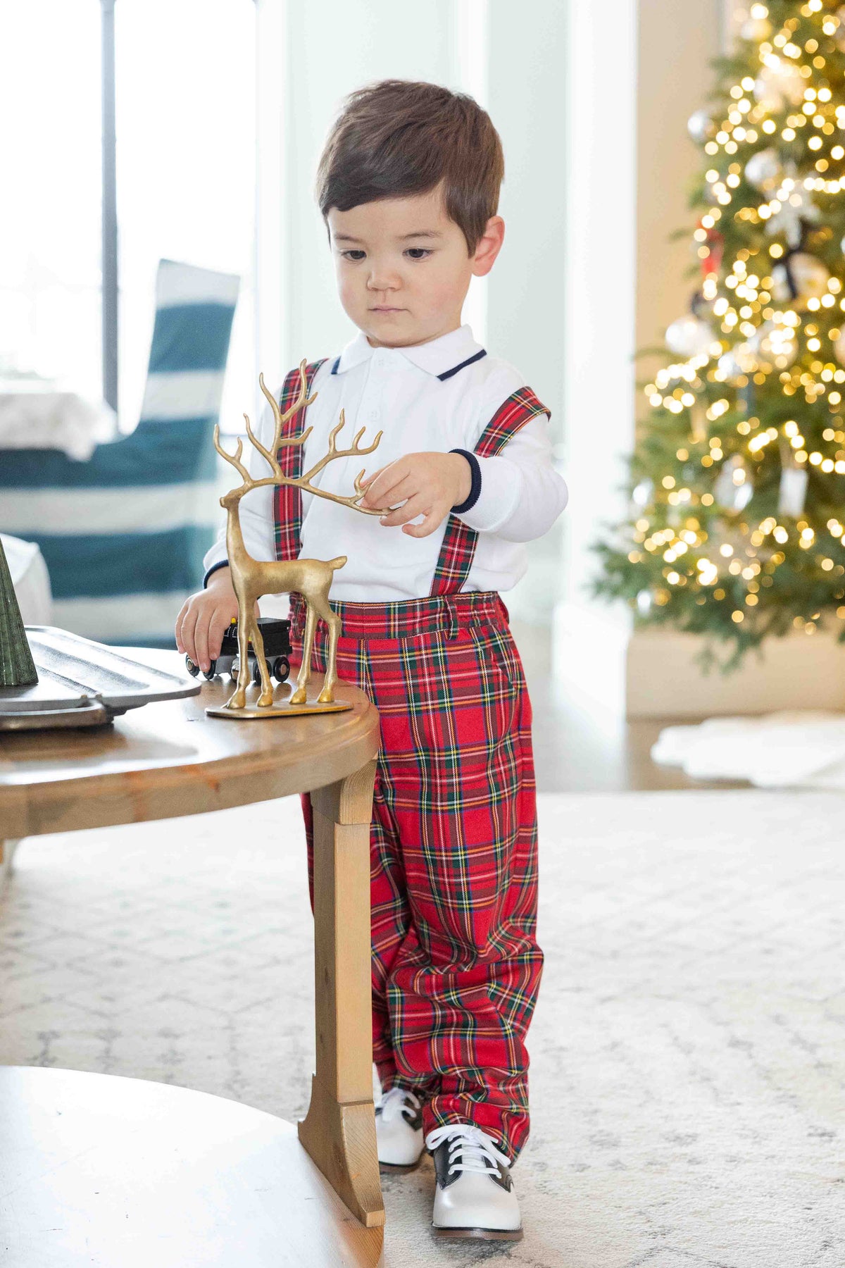 A toddler boy wearing a white long-sleeve shirt and red plaid suspenders with matching plaid pants, standing indoors near a wooden table and a lit Christmas tree in the background.