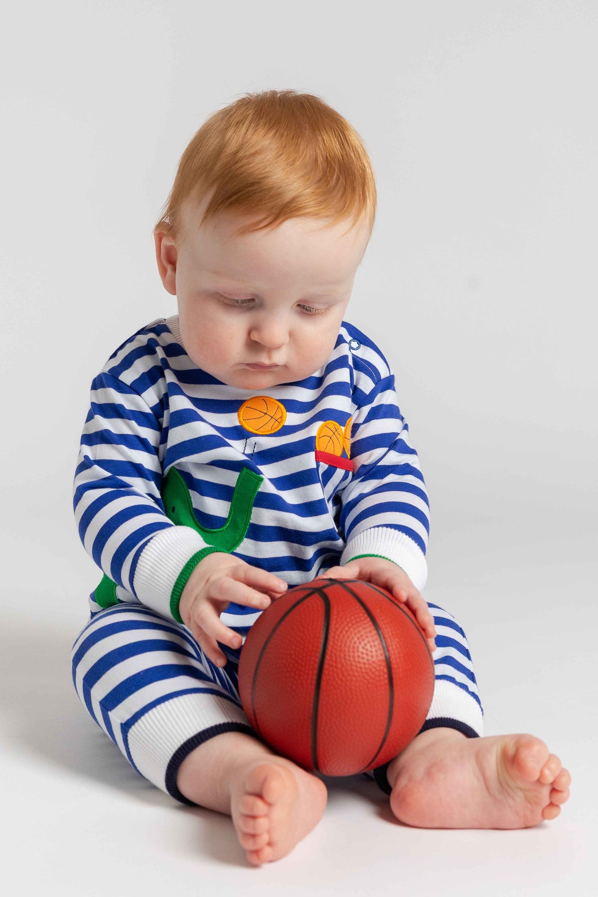 Baby in a blue and white striped outfit holding a red basketball on a light gray background
