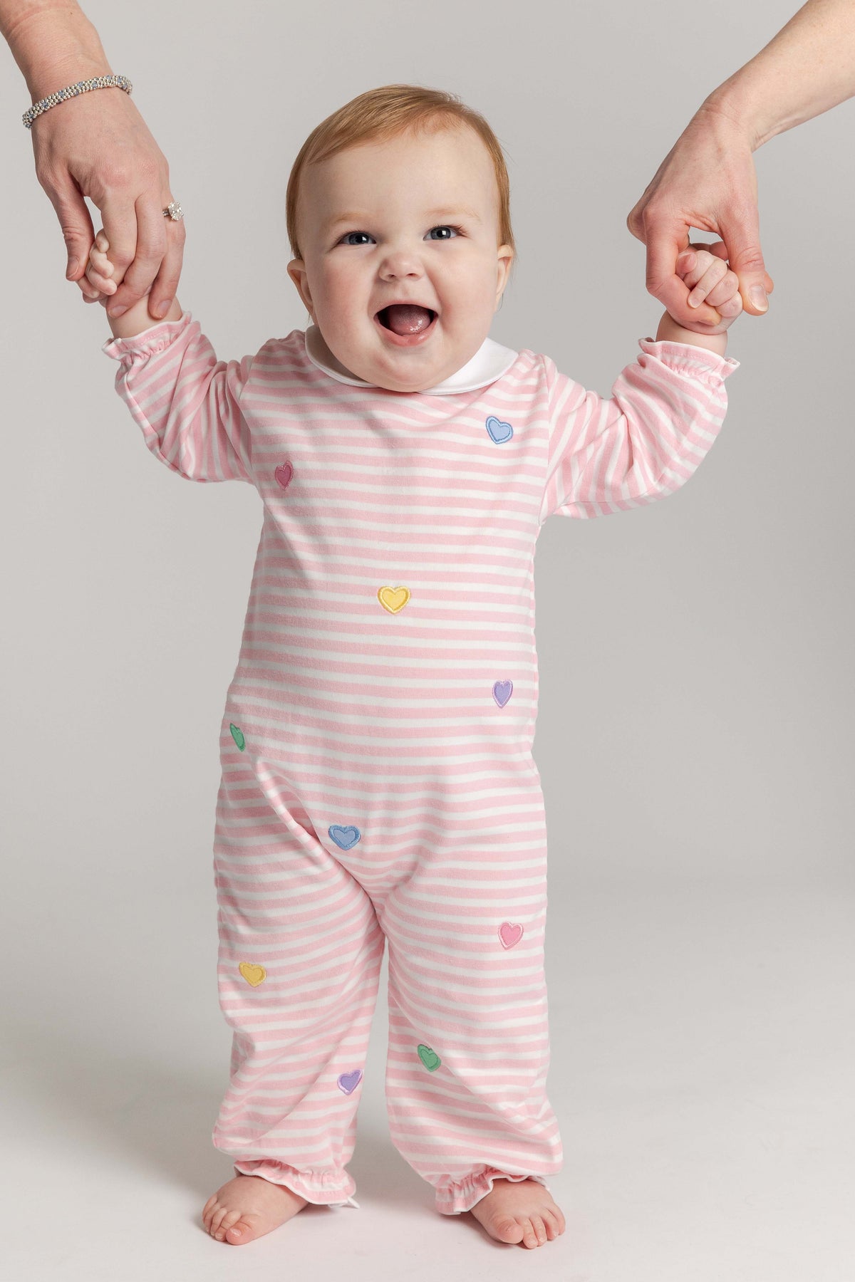 Baby in pink striped pajamas with colorful hearts, standing between two adult hands on a plain background