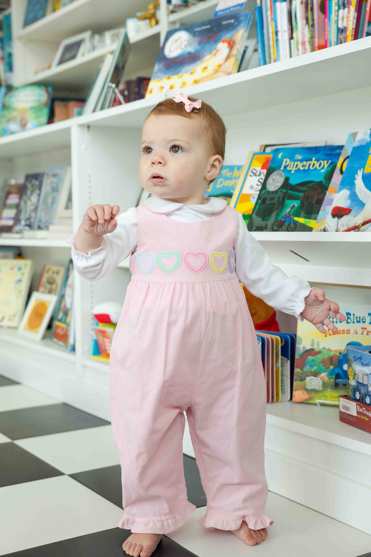 Baby in a pink outfit with heart designs standing in front of a bookshelf filled with books.