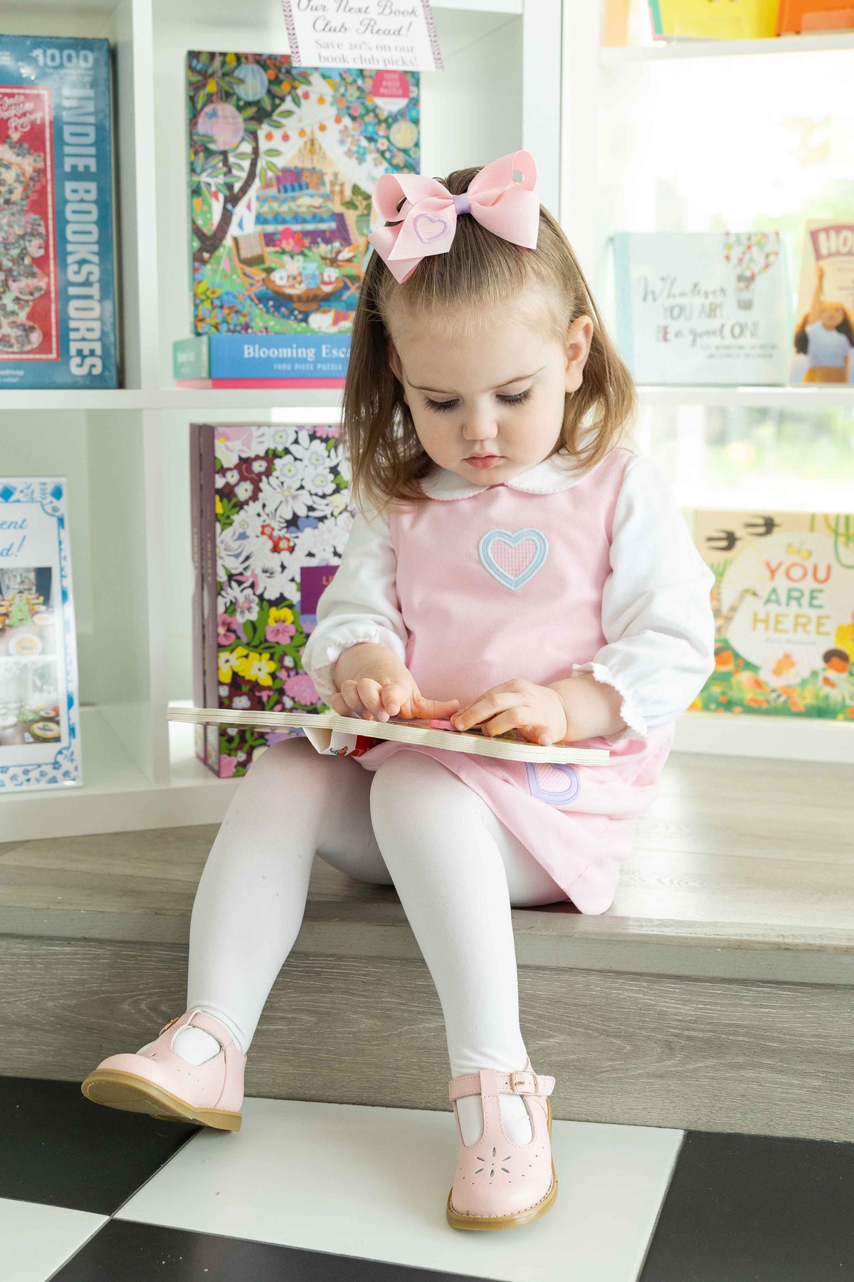 Child in a pink dress sitting on a shelf with books and toys around
