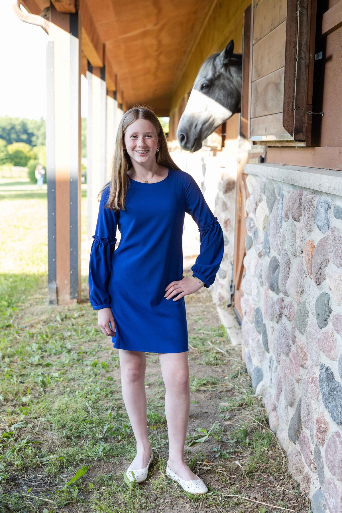 A girl wearing a royal blue long-sleeve dress stands on a farm beside a stone wall and wooden stable, with a horse peeking out from a stall window in the background.