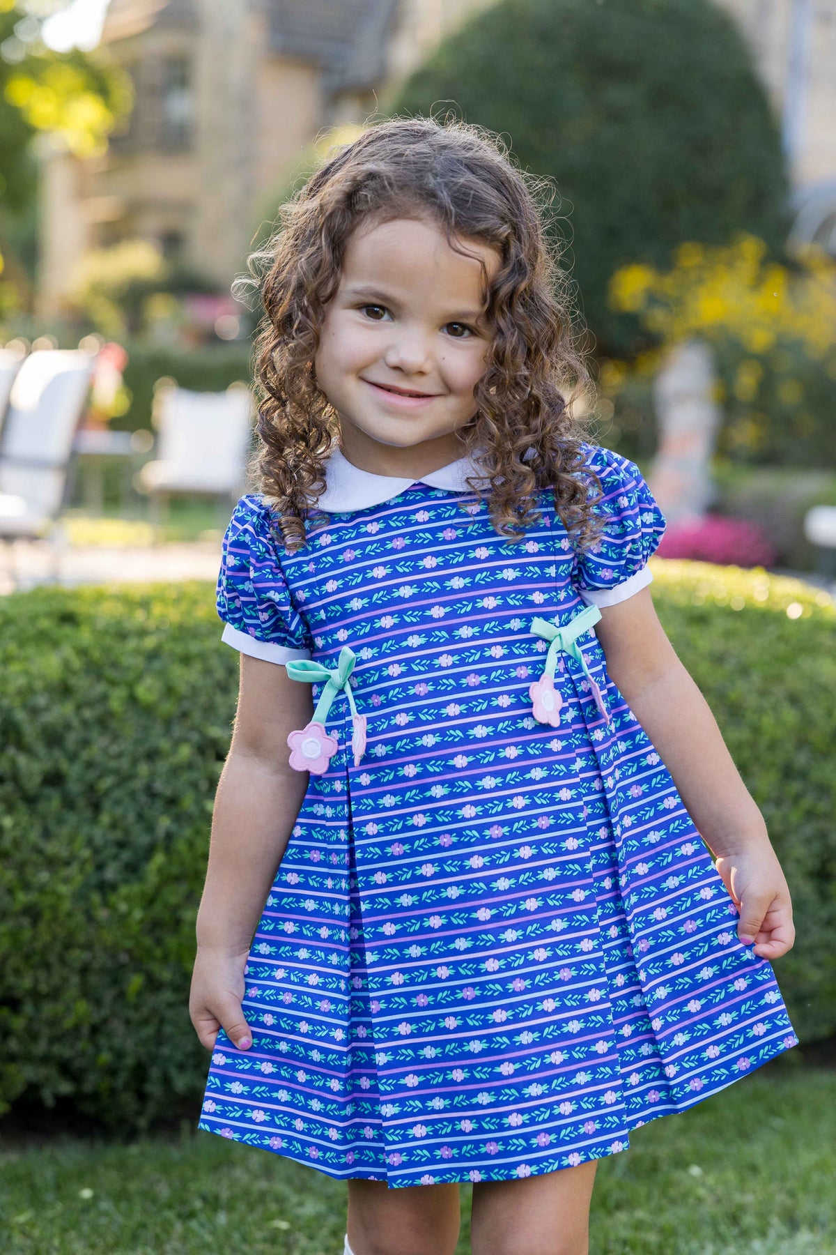 A young girl with curly brown hair stands outdoors in a garden wearing a blue short-sleeve dress with a pattern, a white collar, and mint green ribbon bows with pink flowers.