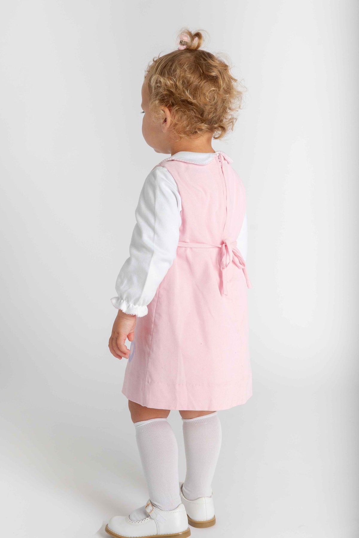 A toddler girl shown from the back wearing a light pink pinafore-style dress over a white long-sleeve blouse, white knee-high socks, and white shoes, standing against a plain light gray studio background.