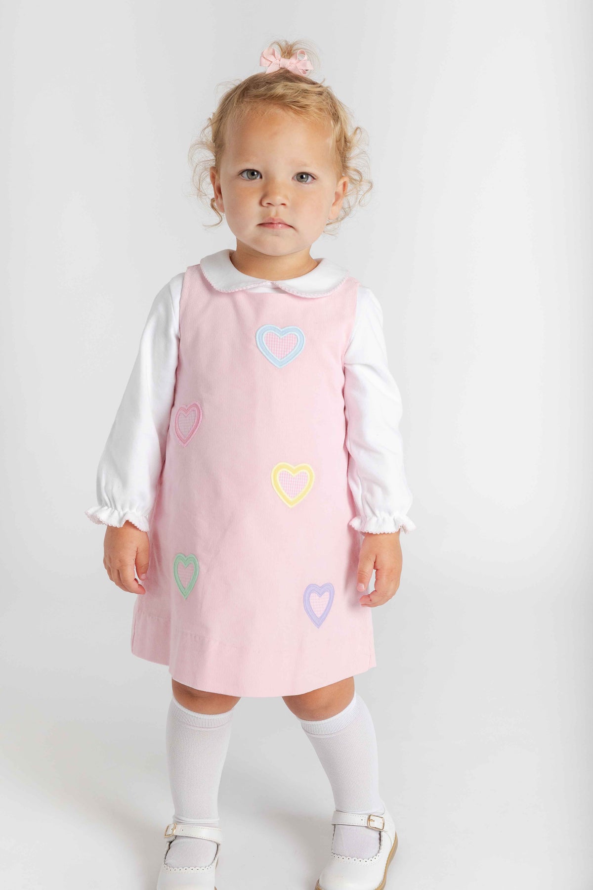 A toddler girl wearing a light pink pinafore dress over a white long-sleeve blouse, white knee-high socks, and white shoes, standing against a plain white studio background.