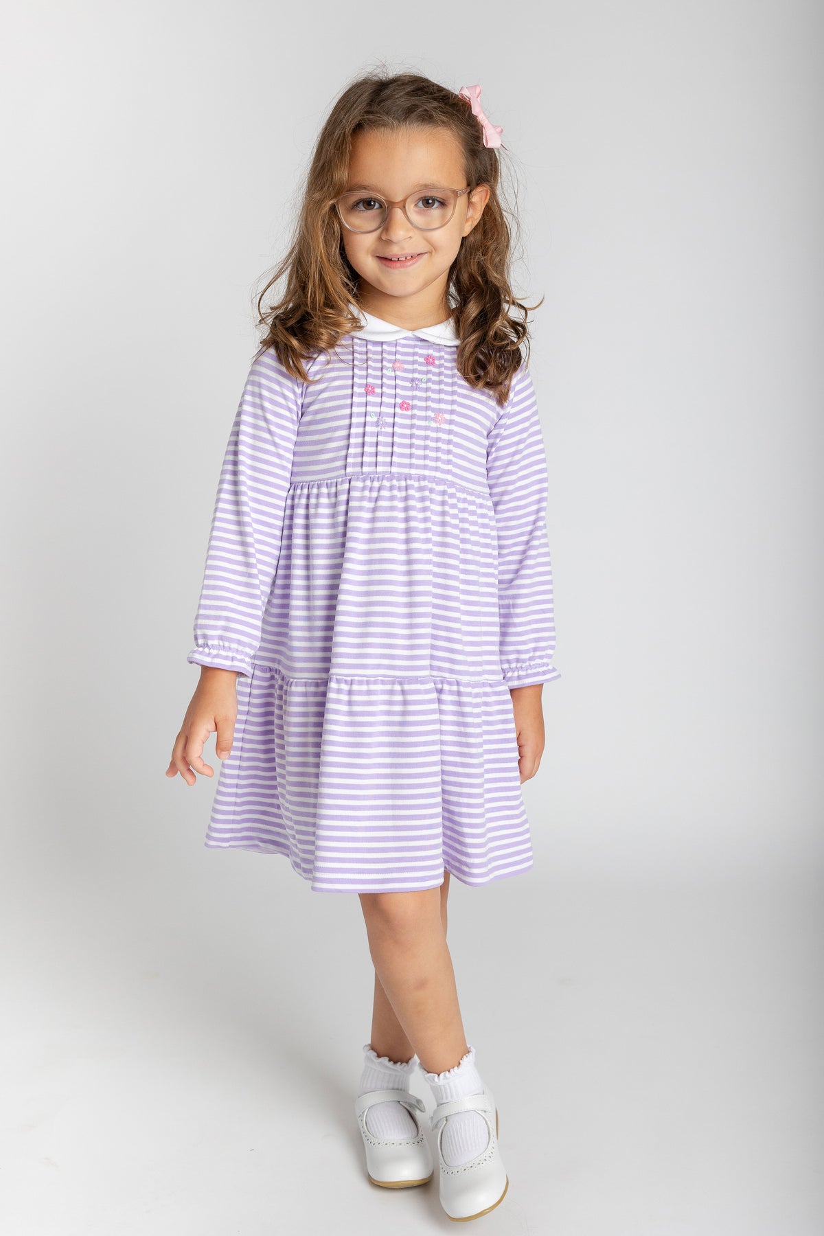 A young girl with glasses and a pink hair clip stands against a light gray studio background, wearing a lavender and white striped tiered dress with white shoes.