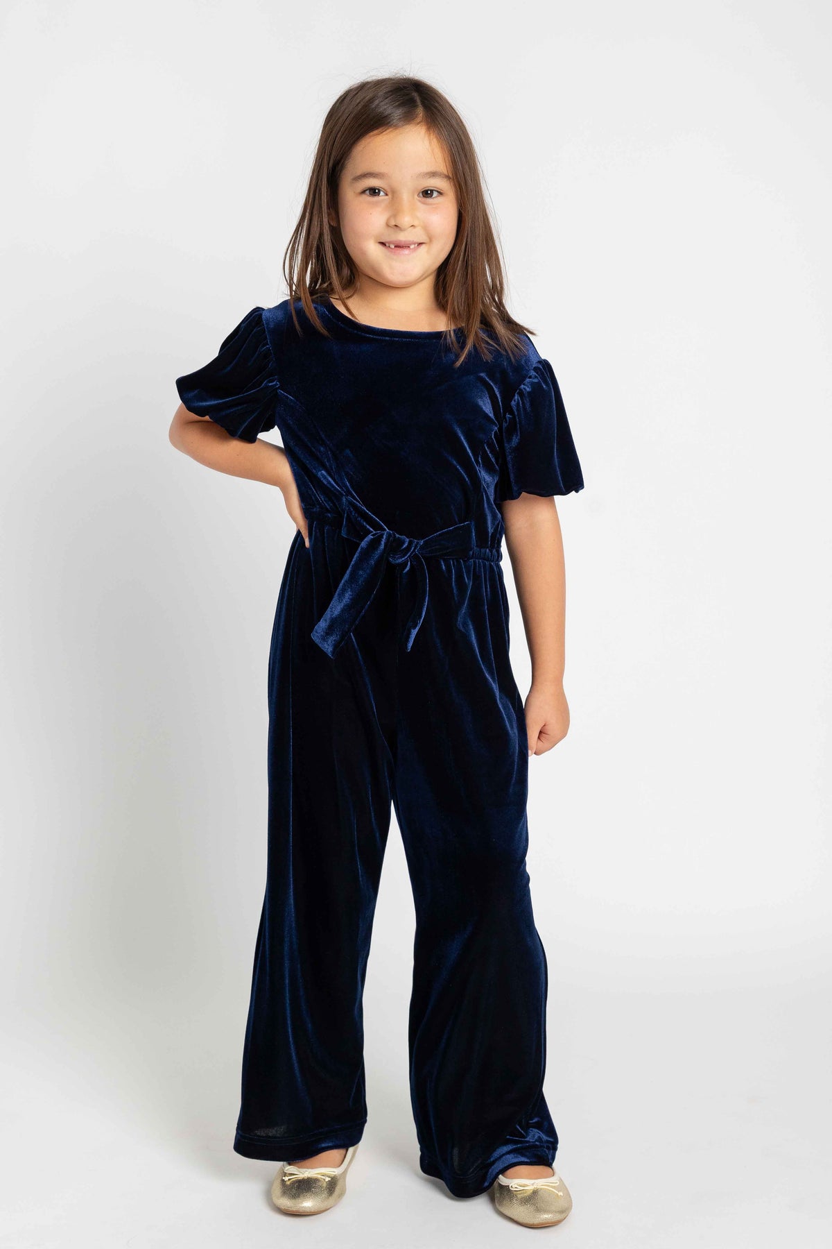 A young girl with long brown hair stands against a light gray studio background wearing a navy blue velvet jumpsuit with short flutter sleeves, a tie waist, and wide legs. She is wearing metallic gold shoes.