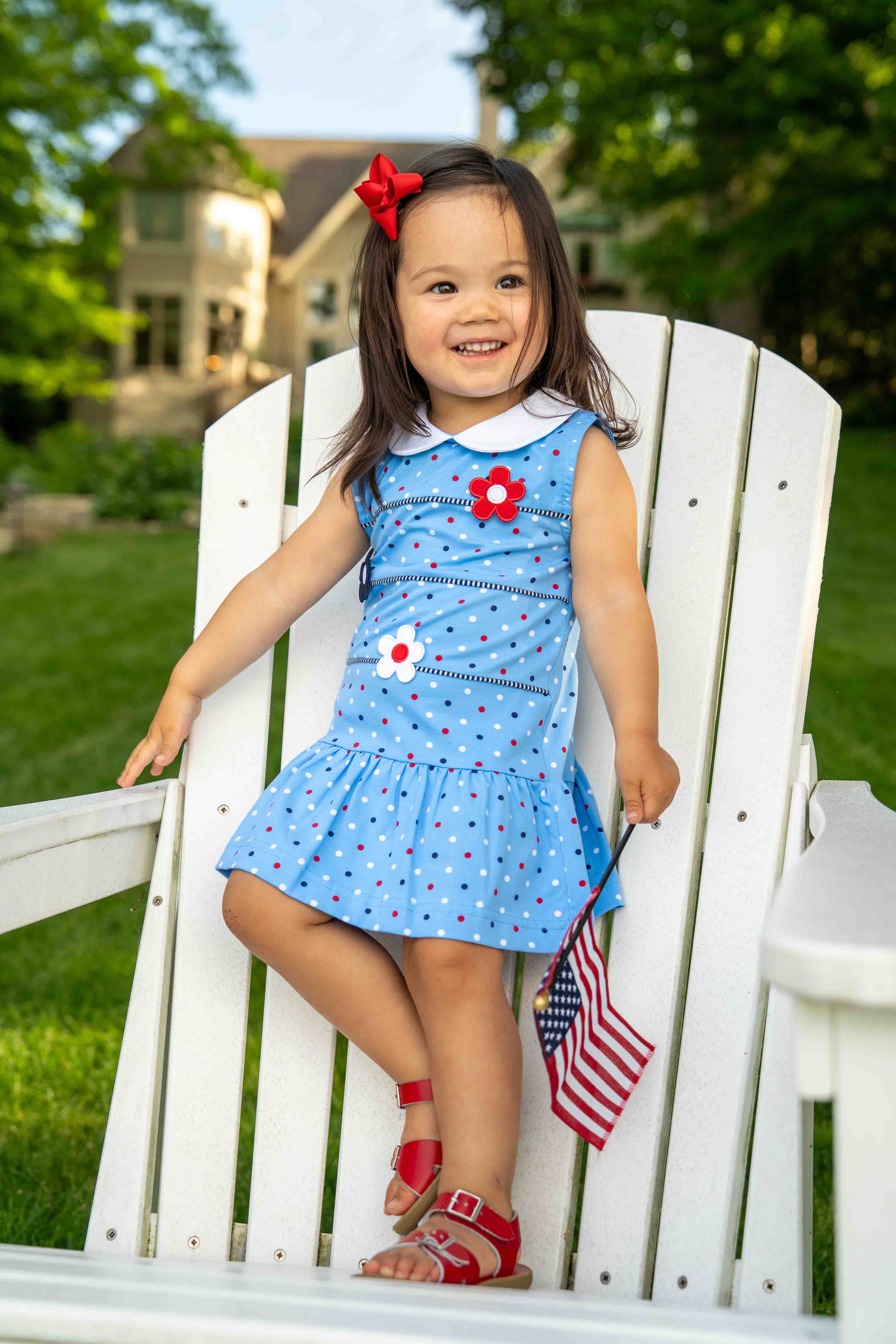 Young girl in a blue dress with a flower design, sitting on a white chair outdoors.