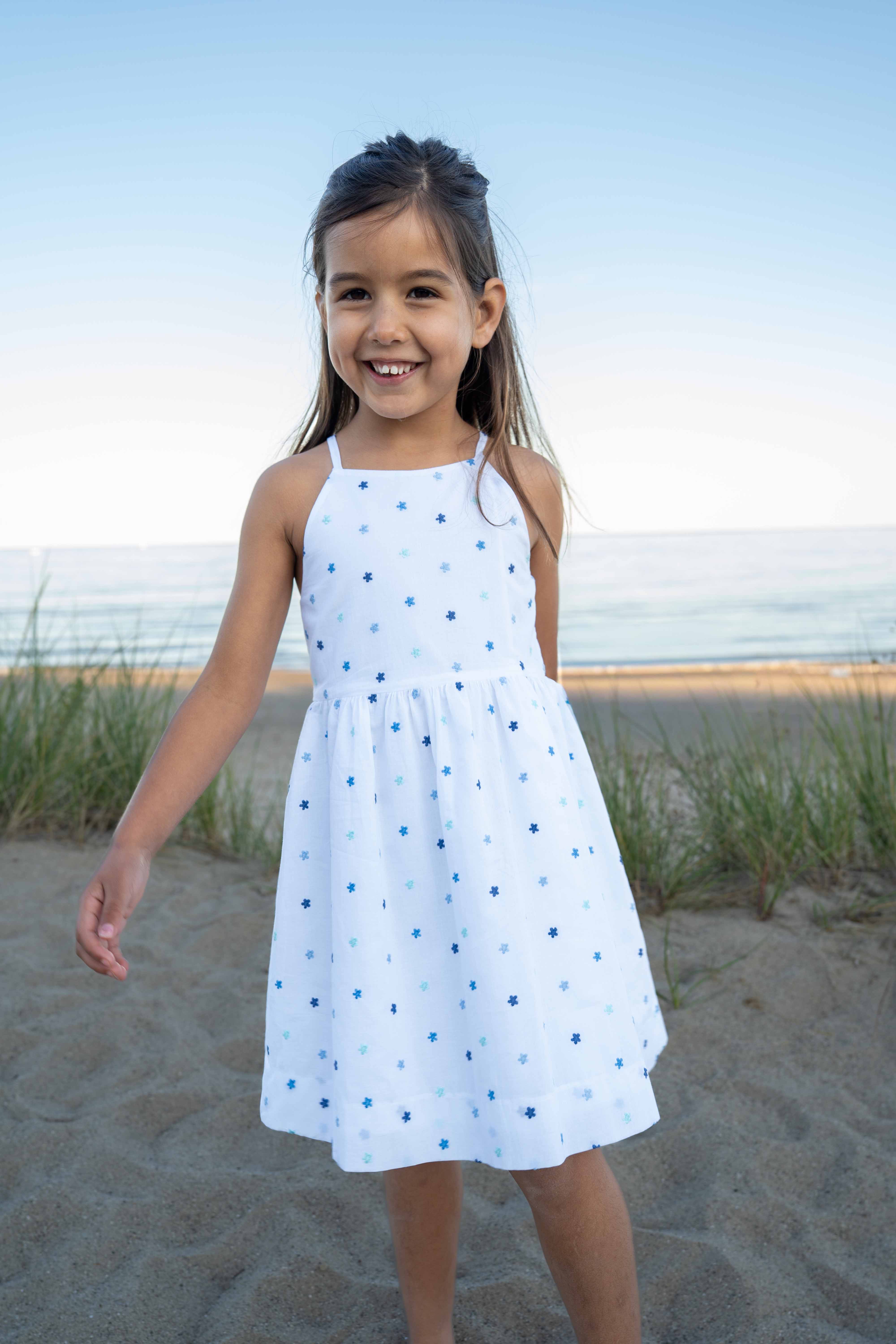 Young girl in a white dress with blue polka dots standing on a sandy beach.