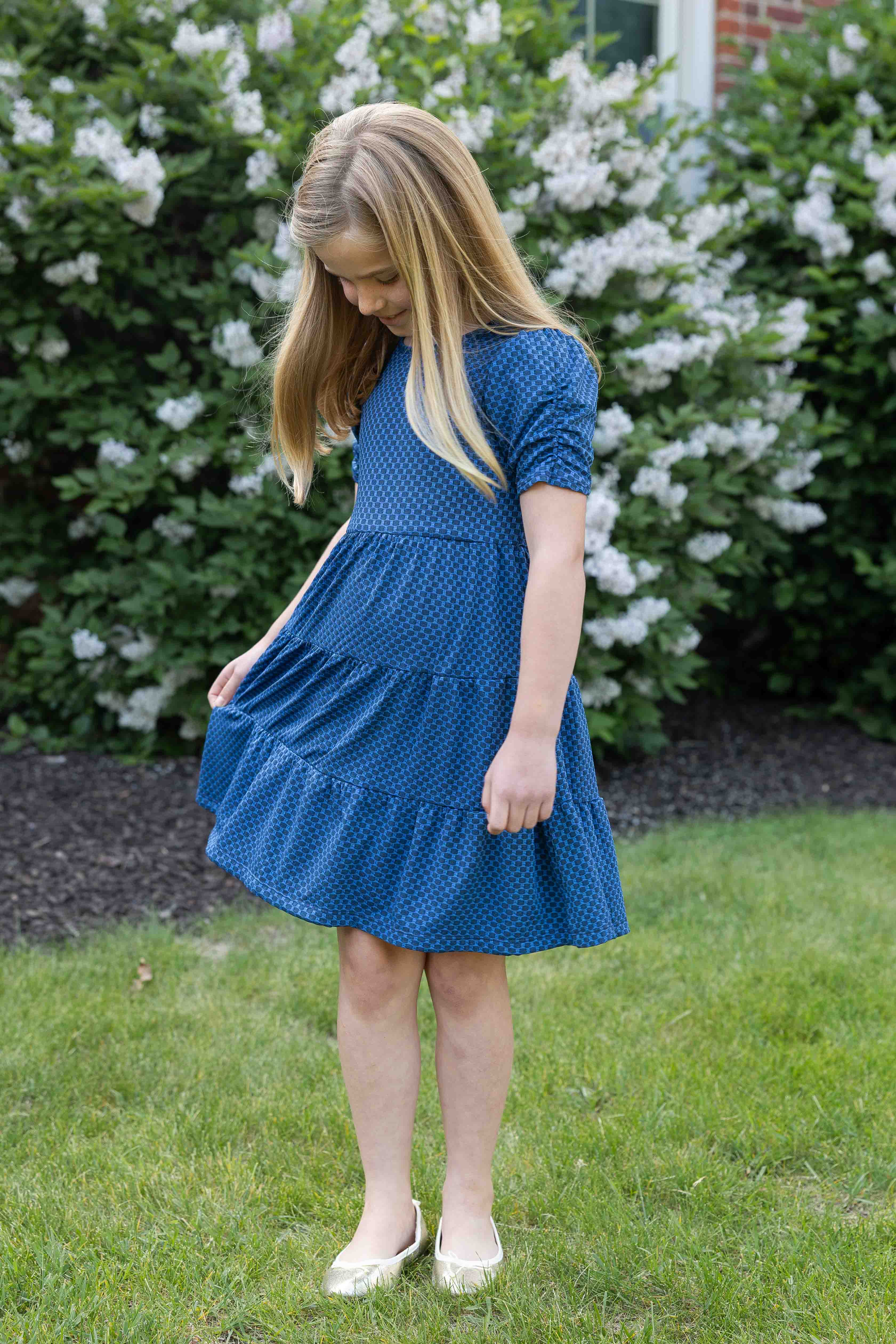 Young girl in a blue dress standing outdoors with greenery in the background