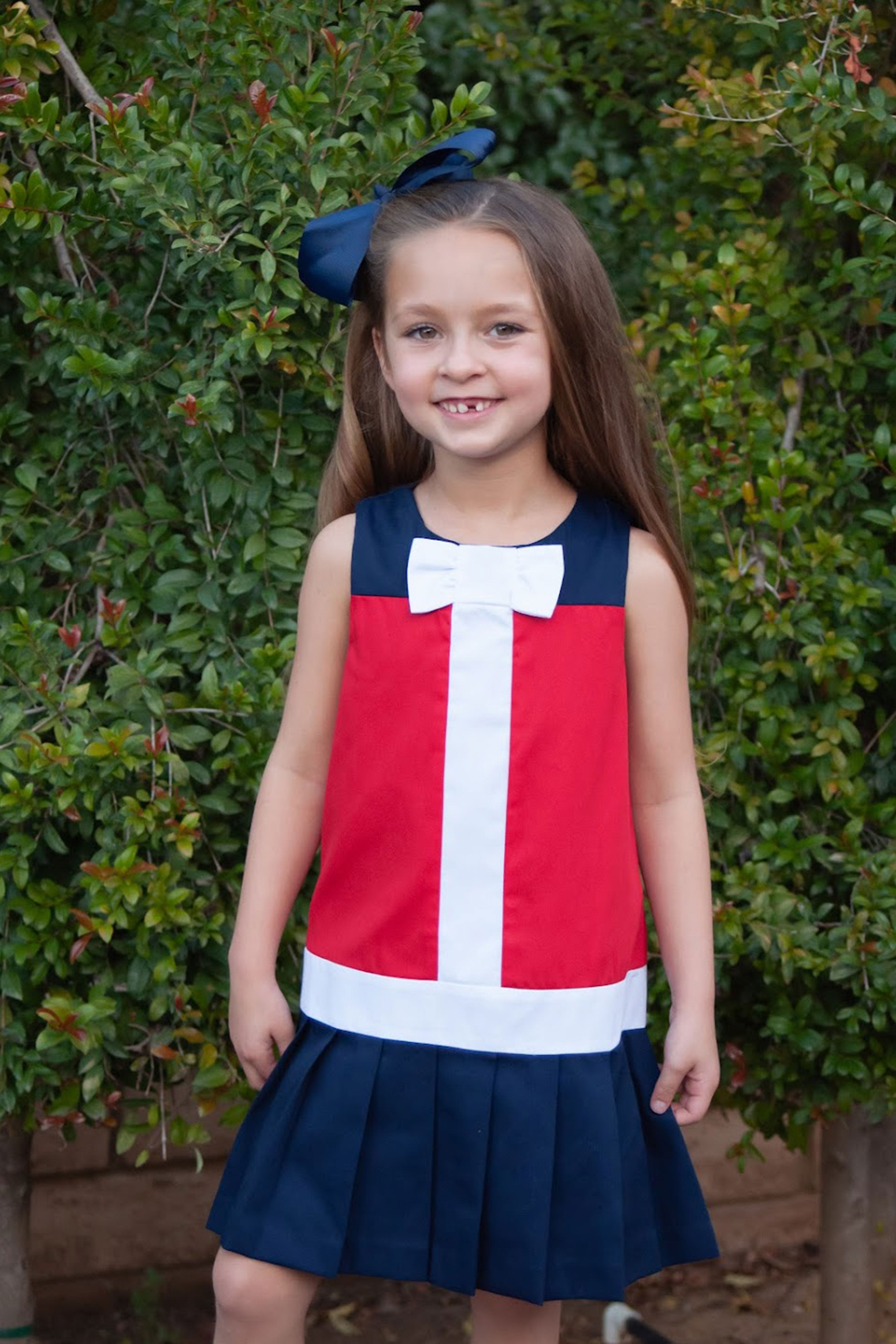 Young girl wearing a red, white, and blue dress with a bow in her hair, standing in front of green bushes.