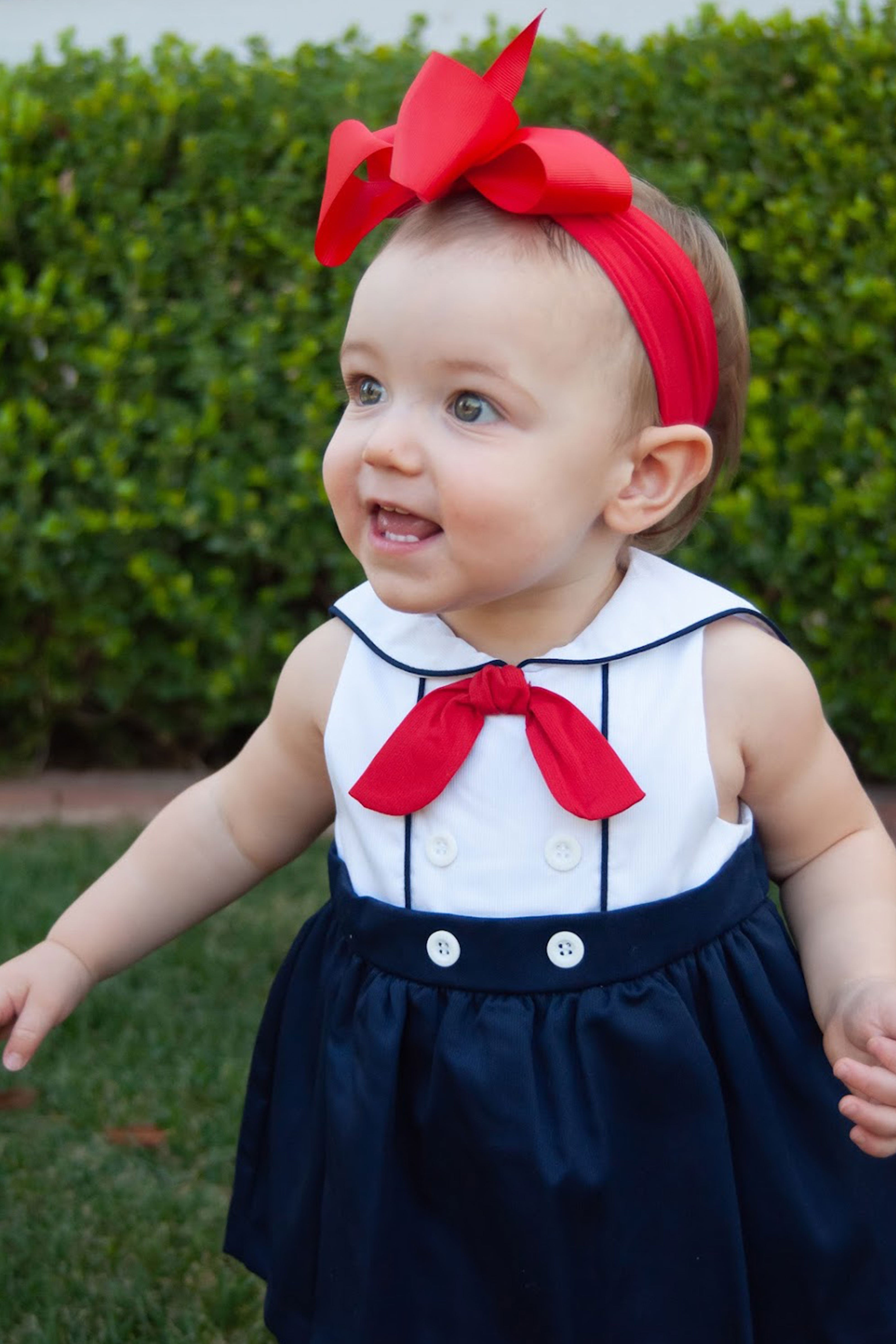 Baby wearing a navy dress with a red bow and headband, standing outdoors.