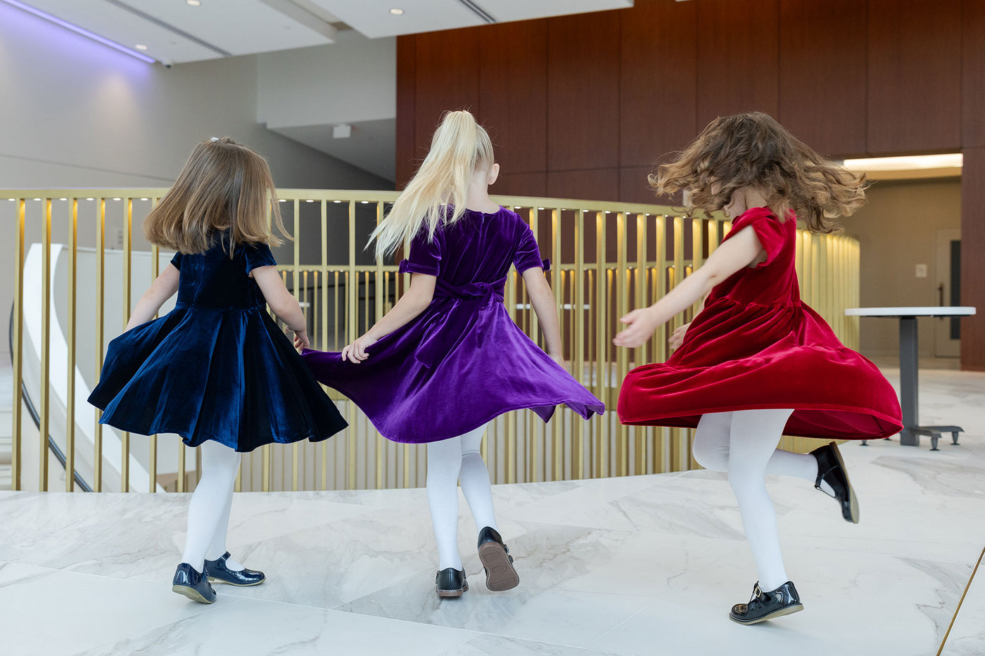 Three children in colorful dresses dancing in a modern indoor setting.
